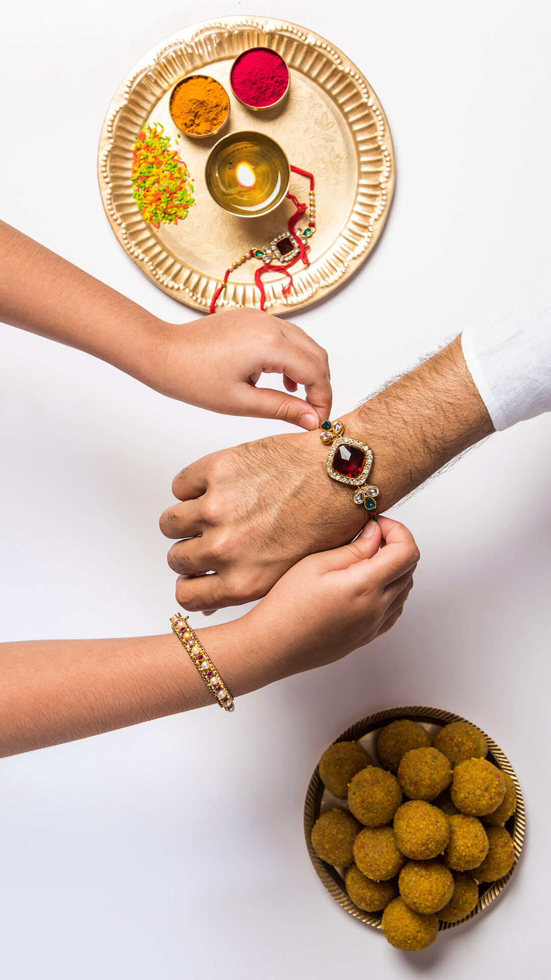 Sister tying an ornate rakhi on her brother’s wrist with traditional sweets, roli, and diya arranged for Raksha Bandhan celebration.
